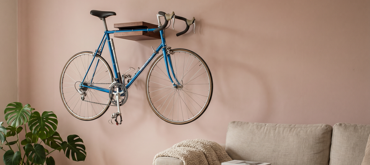 A blue road bicycle with a wooden bike shelf, hanging on a dusty pink wall in a modern living room setting with a sofa, plant, and books.

