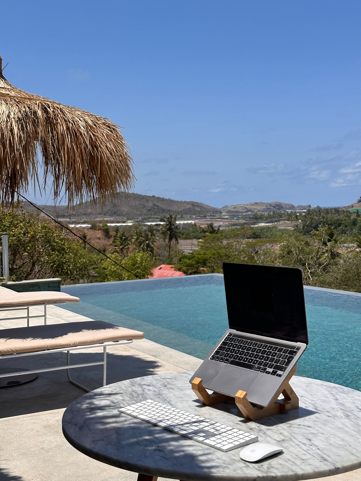 A laptop on a bambooo wooden stand on a round stone table next to an infinity pool.