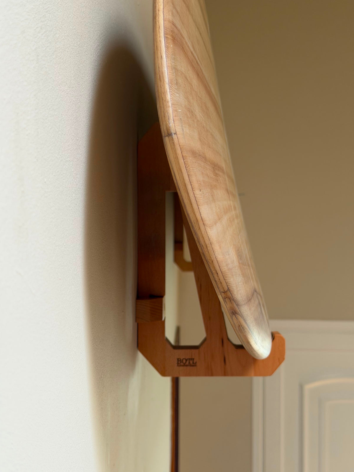 A close-up of a light surfboard mounted on a wooden wall hook, shown against a neutral beige wall.